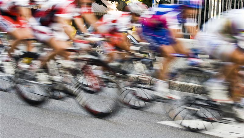 Hình ảnh các vận động viên đua xe đạp ở tốc độ cao trên đường phố, áo quần và bánh xe mờ nhòa - biểu tượng cho tốc độ, cạnh tranh và quyết tâm.