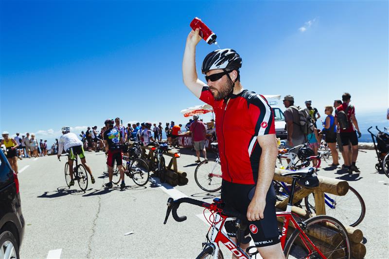 Vận động viên xe đạp đổ nước lên mũ bảo hiểm trong sự kiện ở vùng núi, xung quanh là xe đạp và khán giả.